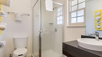 A bathroom with a glass-enclosed shower, a white toilet, a sink with a black countertop, and a mirror with a window above it.