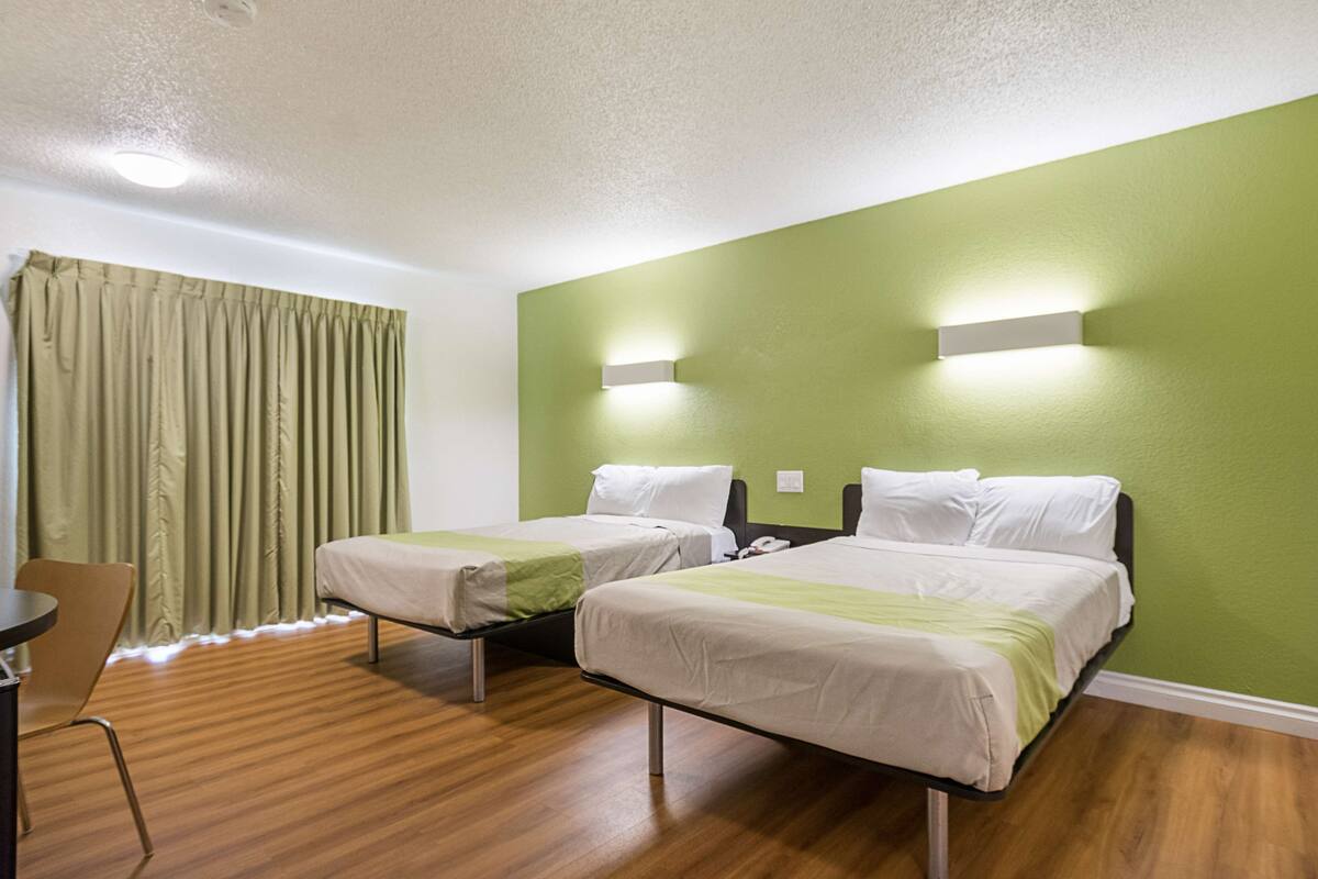 A hotel room with two beds, a wooden floor, a green accent wall, and a window with curtains.