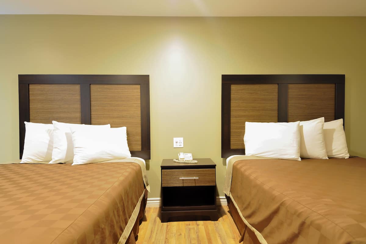 A hotel room with two beds, each with a wooden headboard and white bedding. There is a small bedside table with a lamp and a telephone.