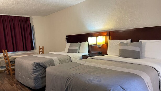 A hotel room with two beds, a wooden chair, a nightstand with a lamp, and a window with curtains.