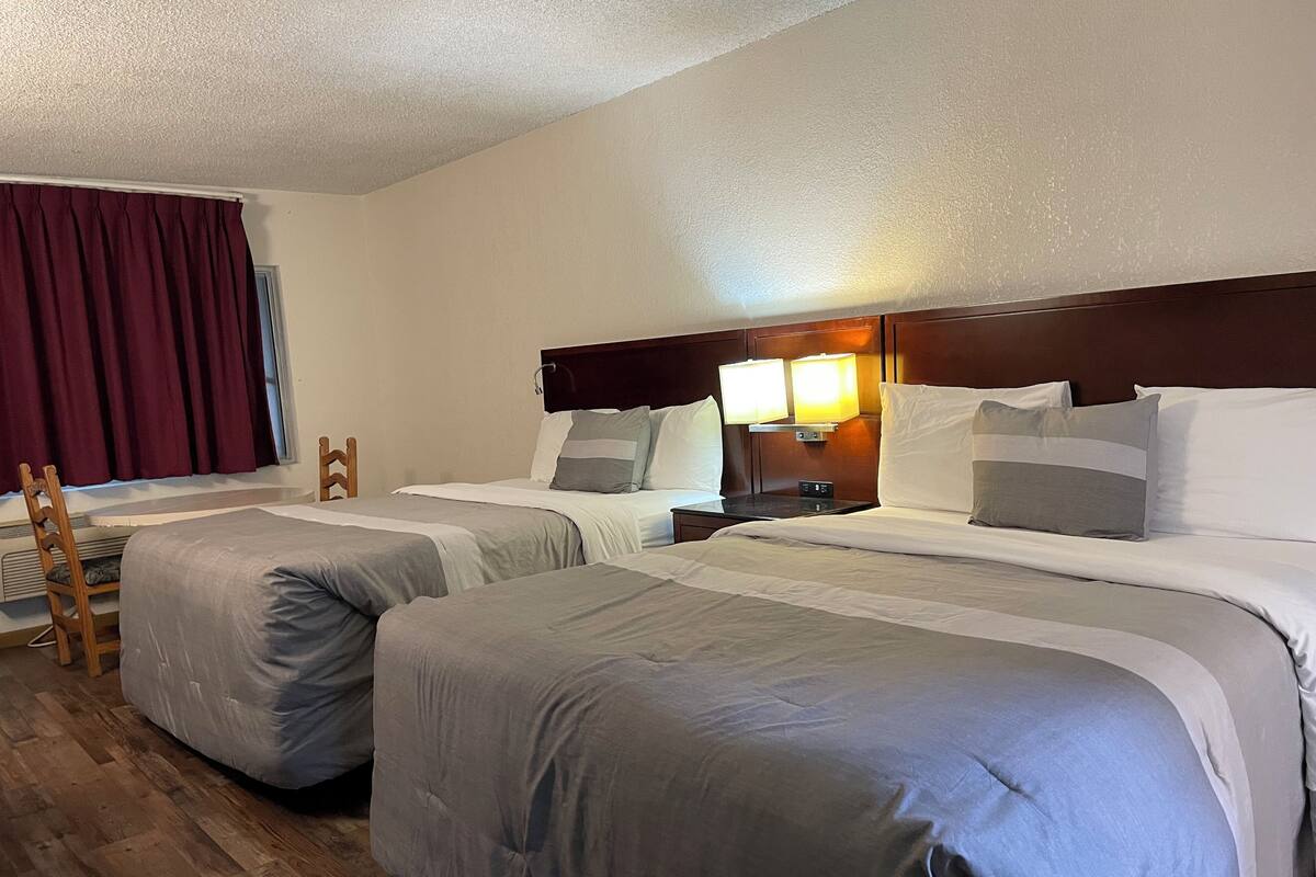 A hotel room with two beds, a wooden chair, a nightstand with a lamp, and a window with curtains.