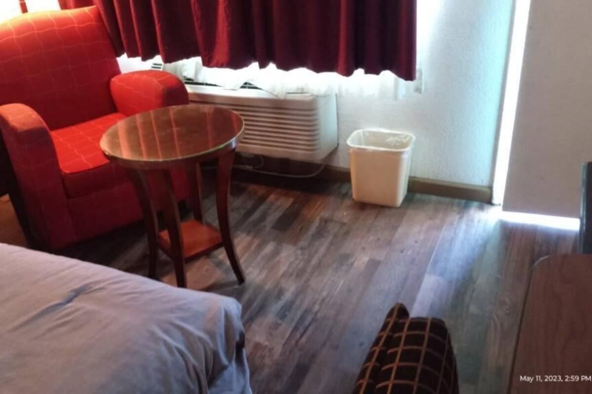 A hotel room with a bed, a red armchair, a small round table, a white trash bin, and a window with curtains.