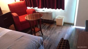 Una habitación de hotel con una cama, un sillón rojo, una mesita redonda pequeña, un cesto de basura blanco y una ventana con cortinas.