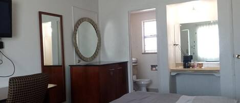 Habitación de hotel con cama, escritorio, silla, armario, espejo, inodoro y ventana.