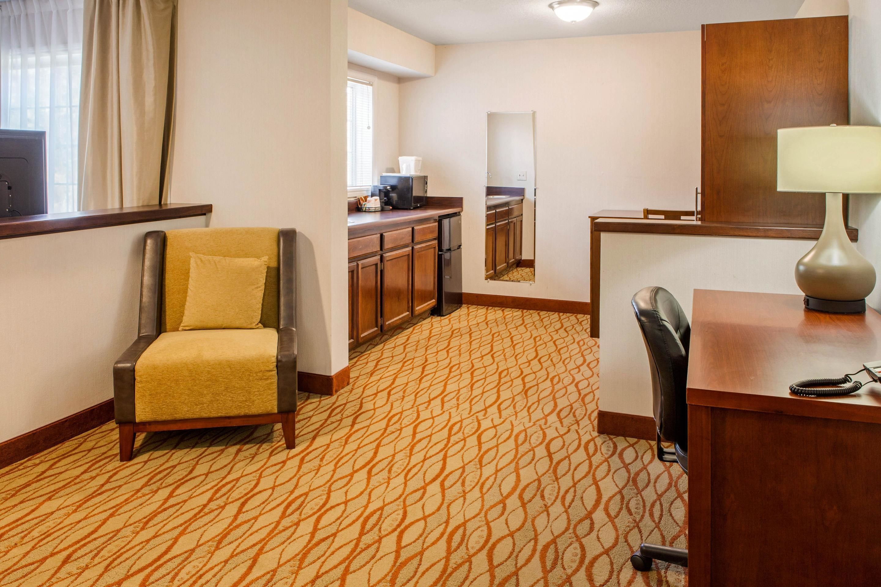 Habitación de hotel con alfombra estampada, un sofá, un escritorio con lámpara, una silla, una zona de cocina con refrigerador y un espejo grande.