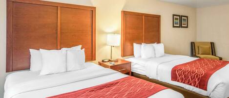 Habitación de hotel con dos camas, cabeceros de madera, ropa de cama blanca, una manta roja con estampado, una lámpara de noche, una silla y una mesita con un teléfono y un libro.