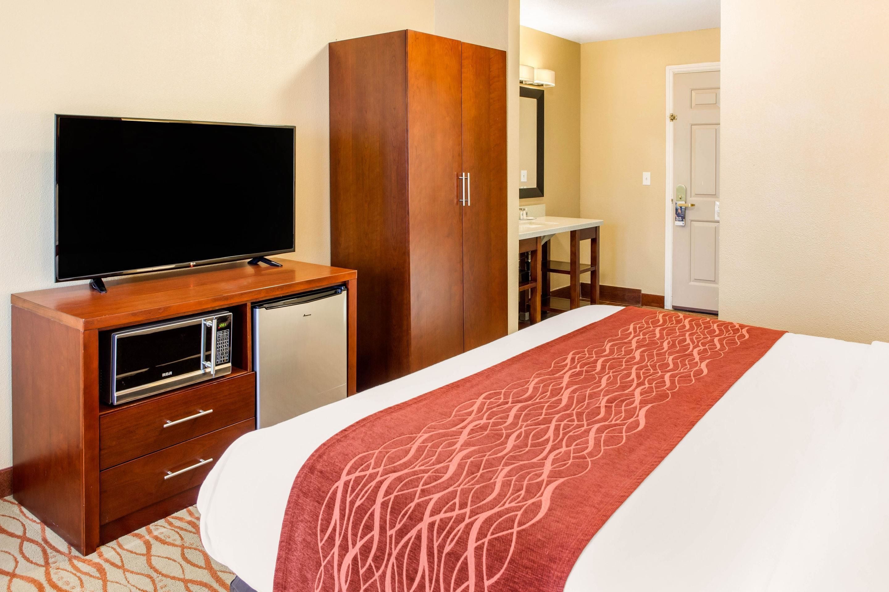 A hotel room with a bed, a TV on a wooden cabinet, a microwave, a small desk, and a mirror.