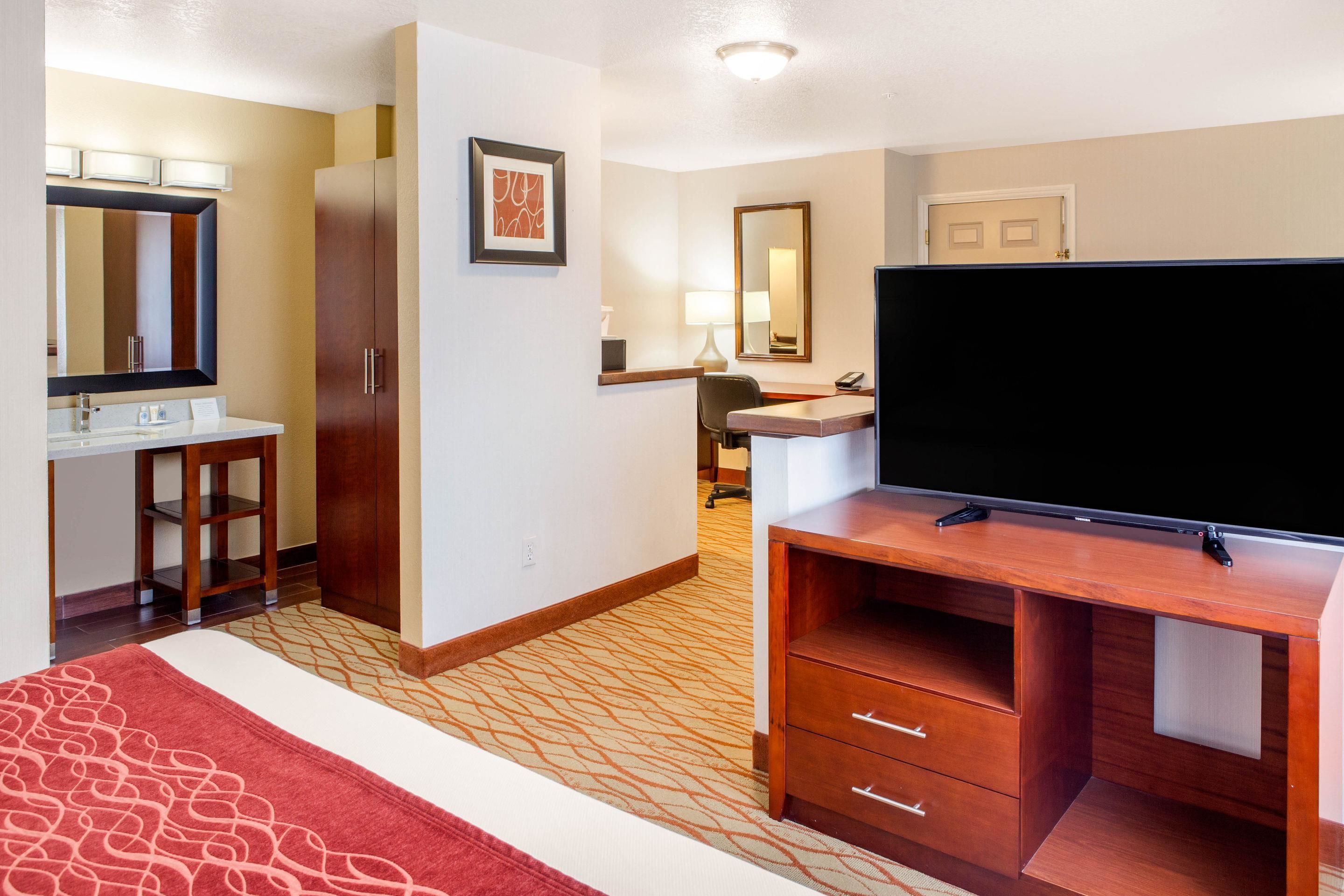 A hotel room with a large flat-screen TV, a wooden desk, a chair, a small stool, a desk lamp, and a large mirror.