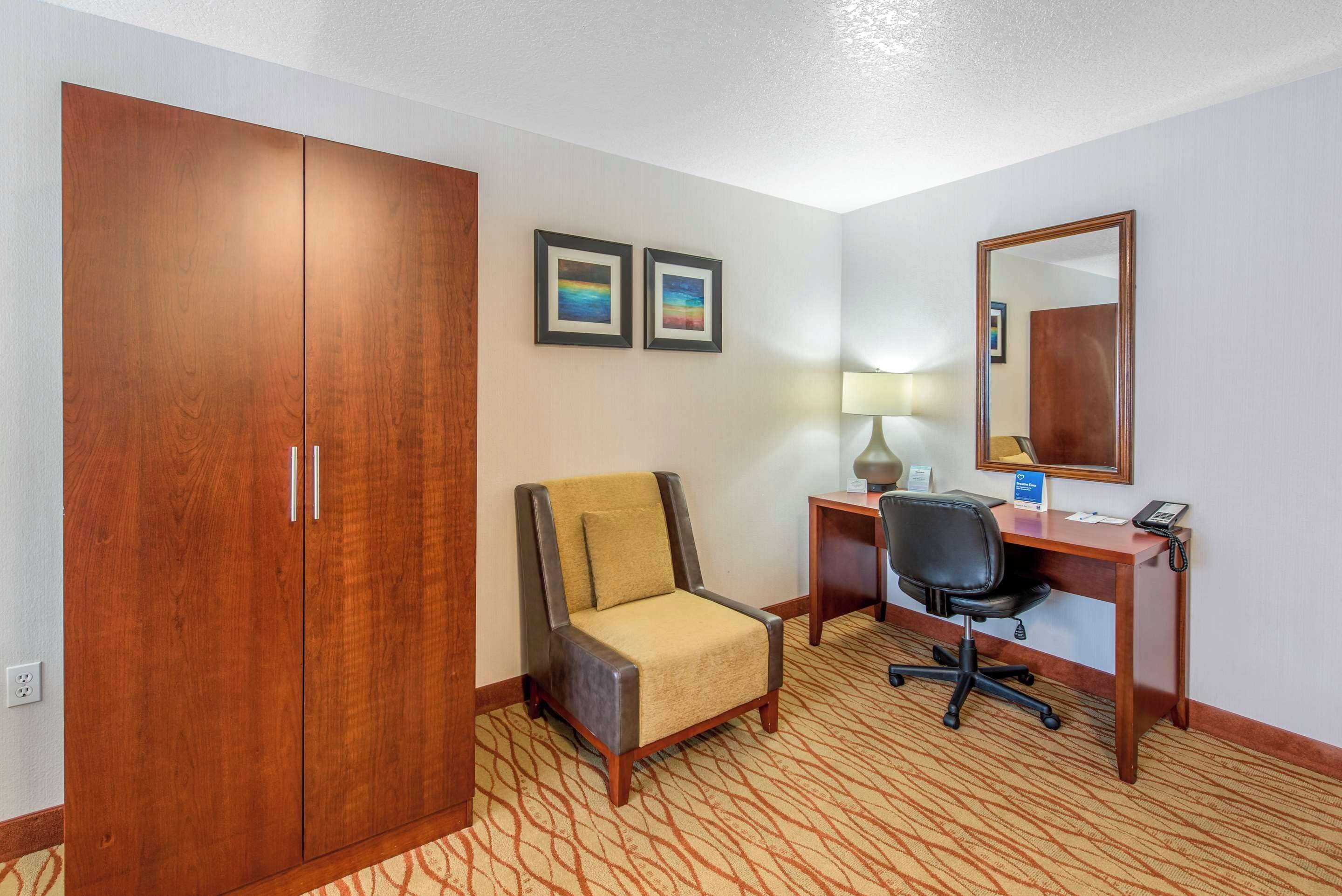 A hotel room with a wooden wardrobe, a cushioned armchair, a desk with a lamp, and a mirror.