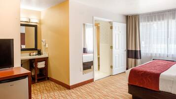 Habitación de hotel con una cama, un escritorio con televisor, un baño con espejo y una puerta que da a otra habitación.