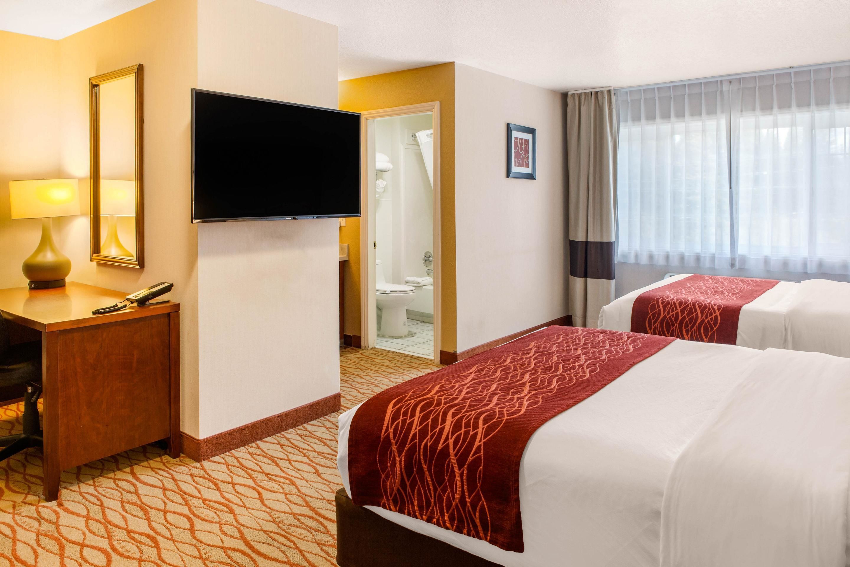 A hotel room with two beds, a desk with a lamp, a TV, and a bathroom visible through an open door.