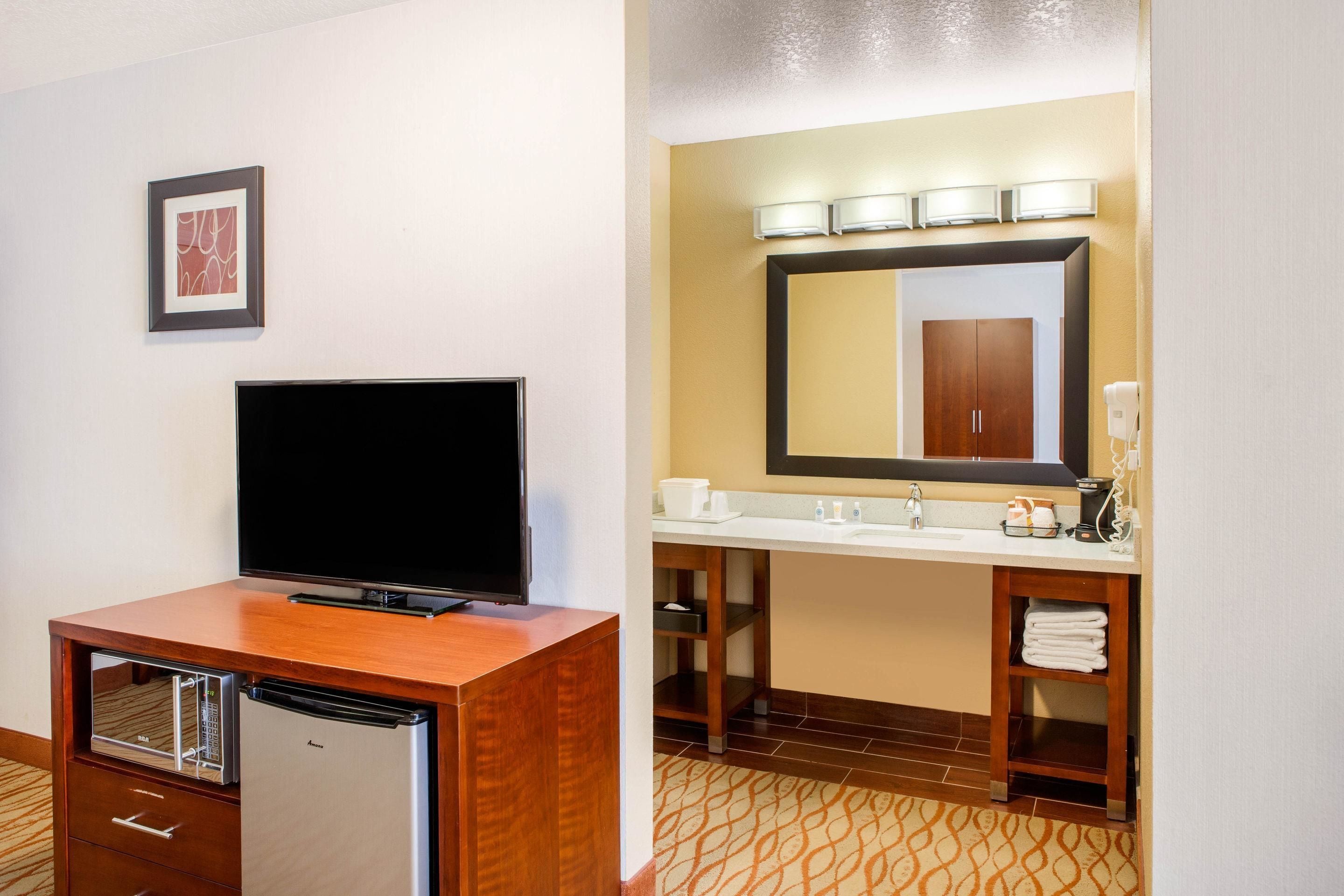 A hotel room with a wooden desk, a flat-screen TV, a minibar, a large mirror above the sink, and a patterned carpet.