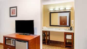 A hotel room with a wooden desk, a flat-screen TV, a minibar, a large mirror above the sink, and a patterned carpet.
