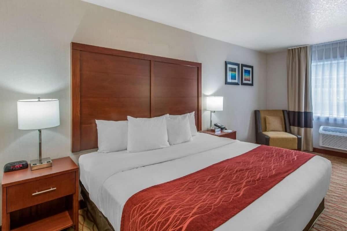 Une chambre d’hôtel avec un grand lit, une table de chevet, une chaise, une lampe et une fenêtre avec des rideaux.