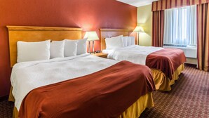 Une chambre d’hôtel avec deux lits, chacun recouvert de draps blancs et d’une couverture rouge. On y trouve des tables de chevet avec des lampes, une fenêtre avec des rideaux et un sol recouvert de moquette.