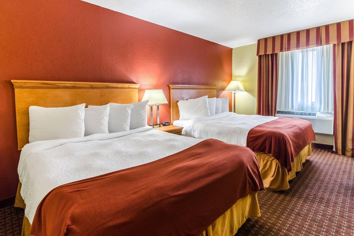 A hotel room with two beds, each with white linens and a red blanket. There are bedside tables with lamps, a window with curtains, and a carpeted floor.