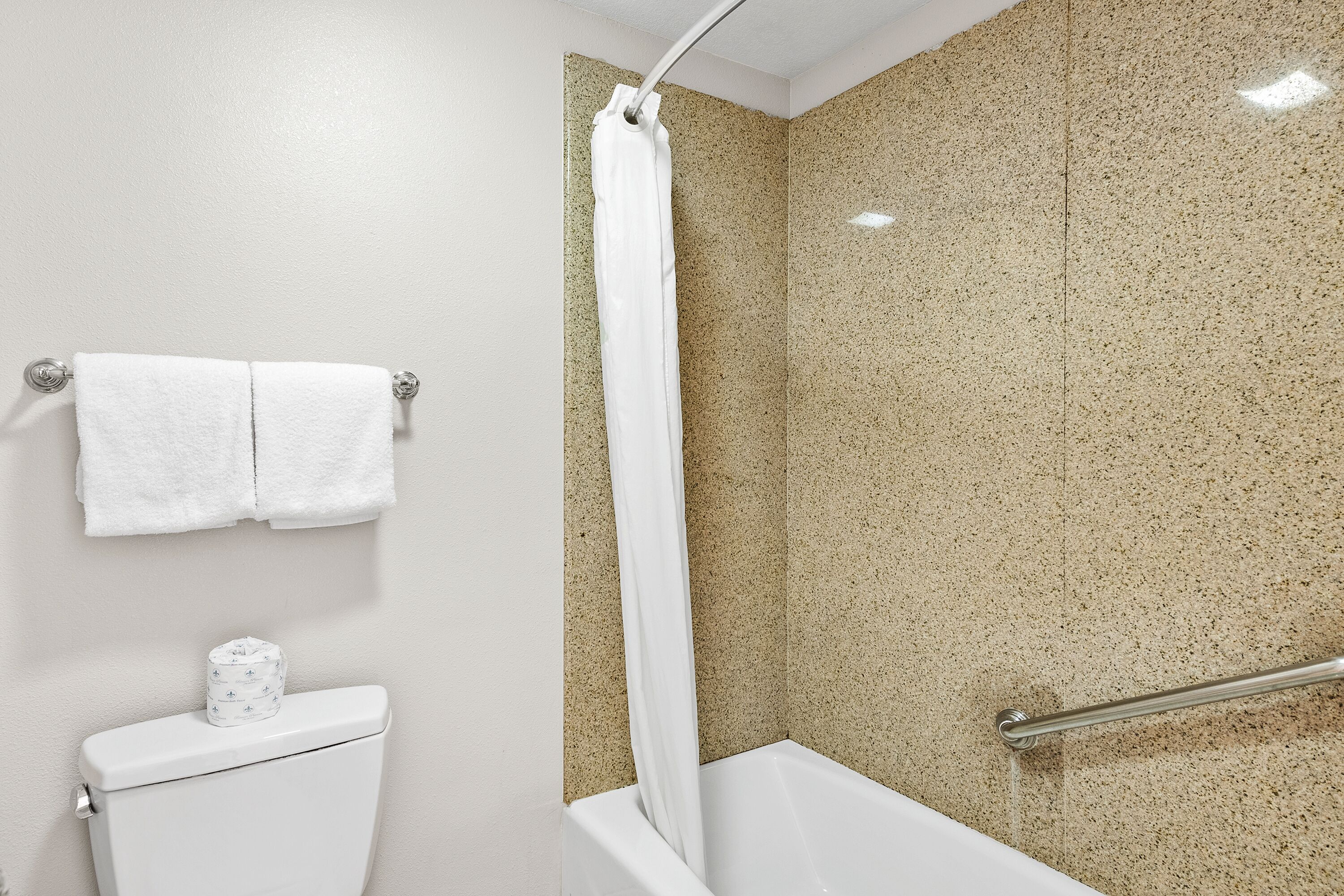 Un baño con inodoro blanco, ducha con pared beige salpicada y toalla blanca colgada en un toallero metálico.