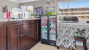 A hotel reception area with a wooden counter, a refrigerator stocked with beverages, a computer monitor, and a large window offering a view of a building and palm trees.