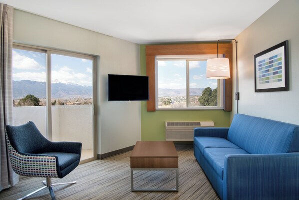 A modern hotel room with a balcony, a flat-screen TV, a blue sofa, a patterned armchair, and a small wooden table. - Holiday Inn Express Colorado Springs Airport by IHG (Colorado Springs)