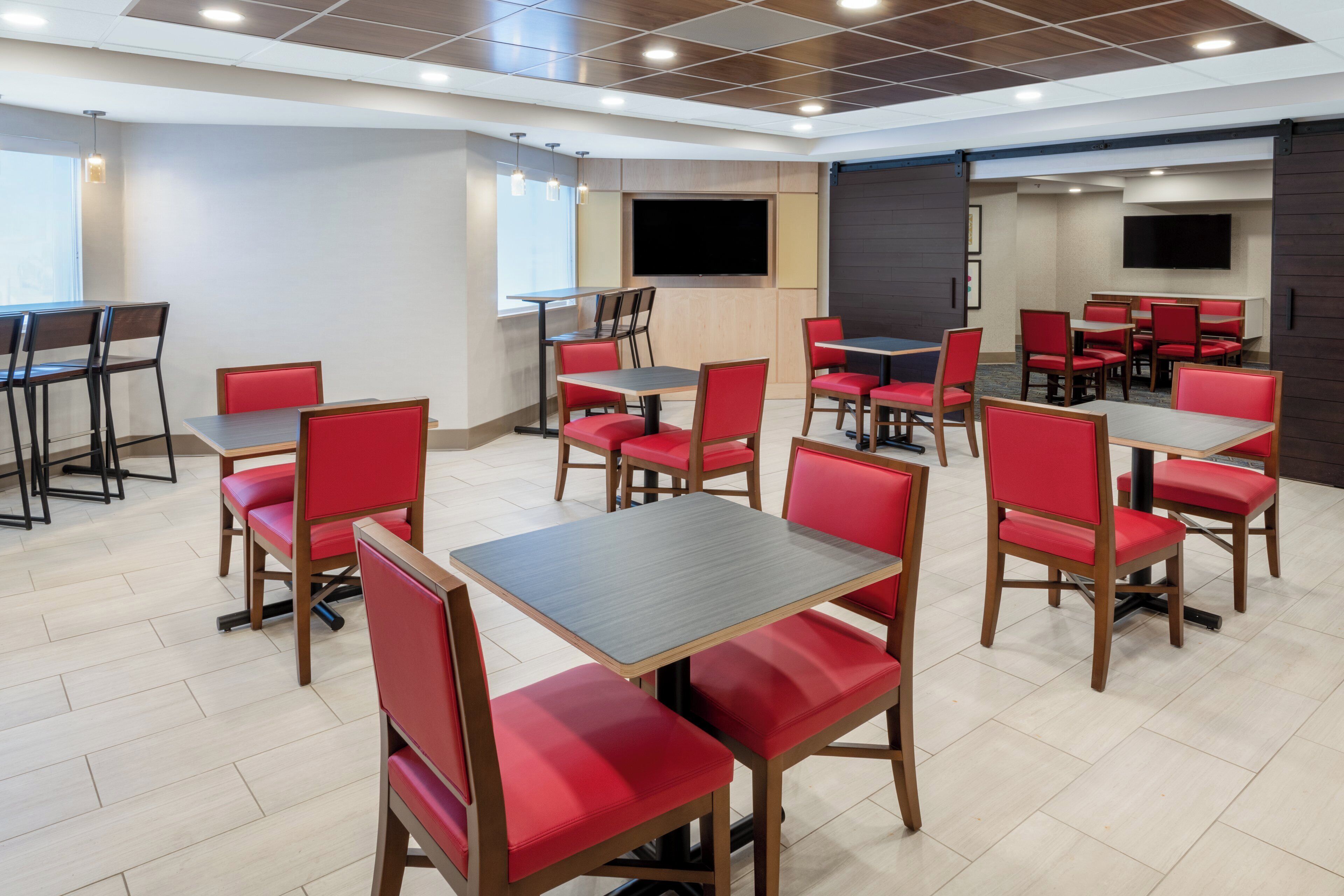 A dining area with multiple tables and chairs, a flat-screen TV mounted on the wall, and a tiled floor.