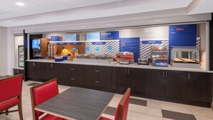 A modern breakfast area with a counter displaying various food options, a seating area with red chairs and a table, and a bakery section. - Holiday Inn Express Colorado Springs Airport by IHG (Colorado Springs)