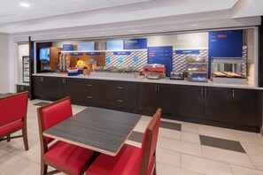 Un espace moderne dédié au petit-déjeuner, avec un comptoir où sont présentés différents choix de plats, un coin salon avec des chaises rouges et une table, ainsi qu’un espace boulangerie.