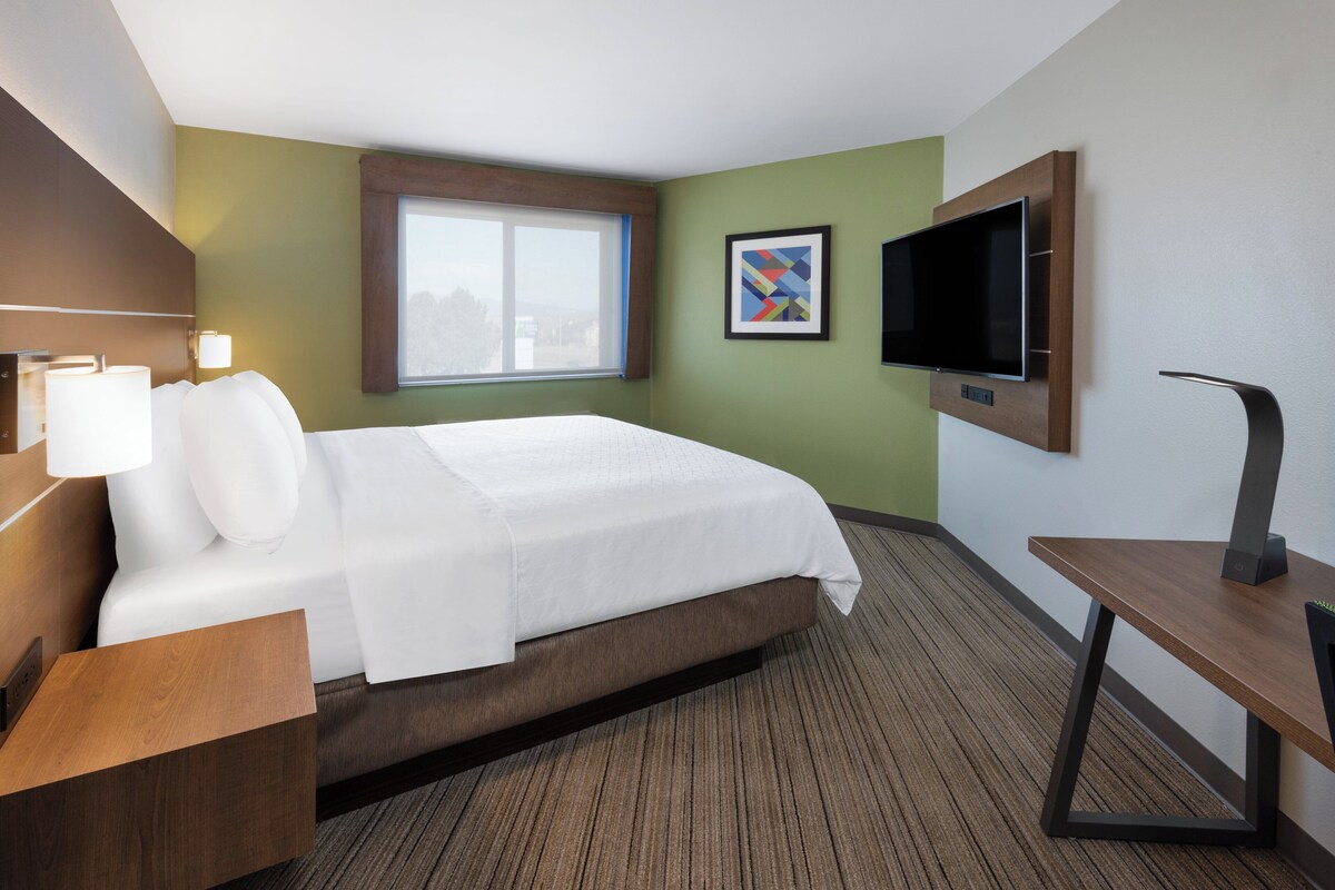 A hotel room with a bed, a TV mounted on the wall, a desk, and a window.