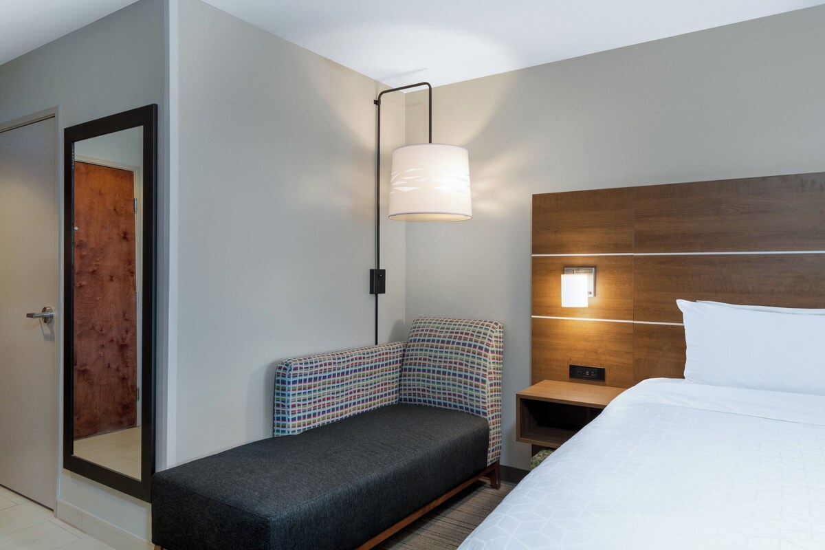 A hotel room with a bed, a small sofa, a nightstand, a wall-mounted lamp, and a wooden headboard.