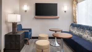 A modern hotel room with a flat-screen TV, a sofa, two ottomans, a lamp, and two small tables. - Hampton Inn Loveland (Loveland)