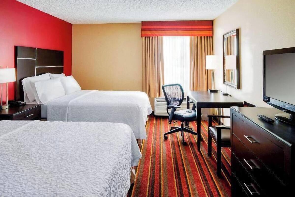 A hotel room with two beds, a desk, a chair, a TV, and a window with curtains.