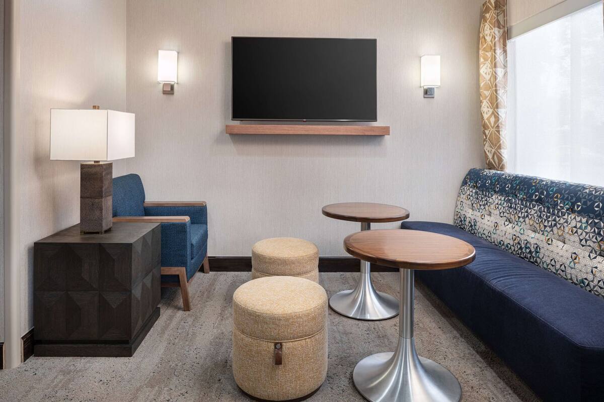 A hotel room with a TV, sofa, two ottomans, a lamp, and two small tables.