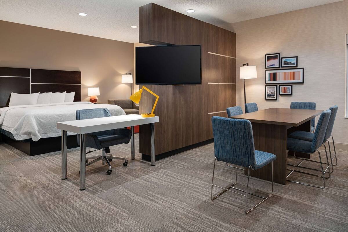A hotel room with a bed, desk, chairs, a TV, and artwork on the wall.