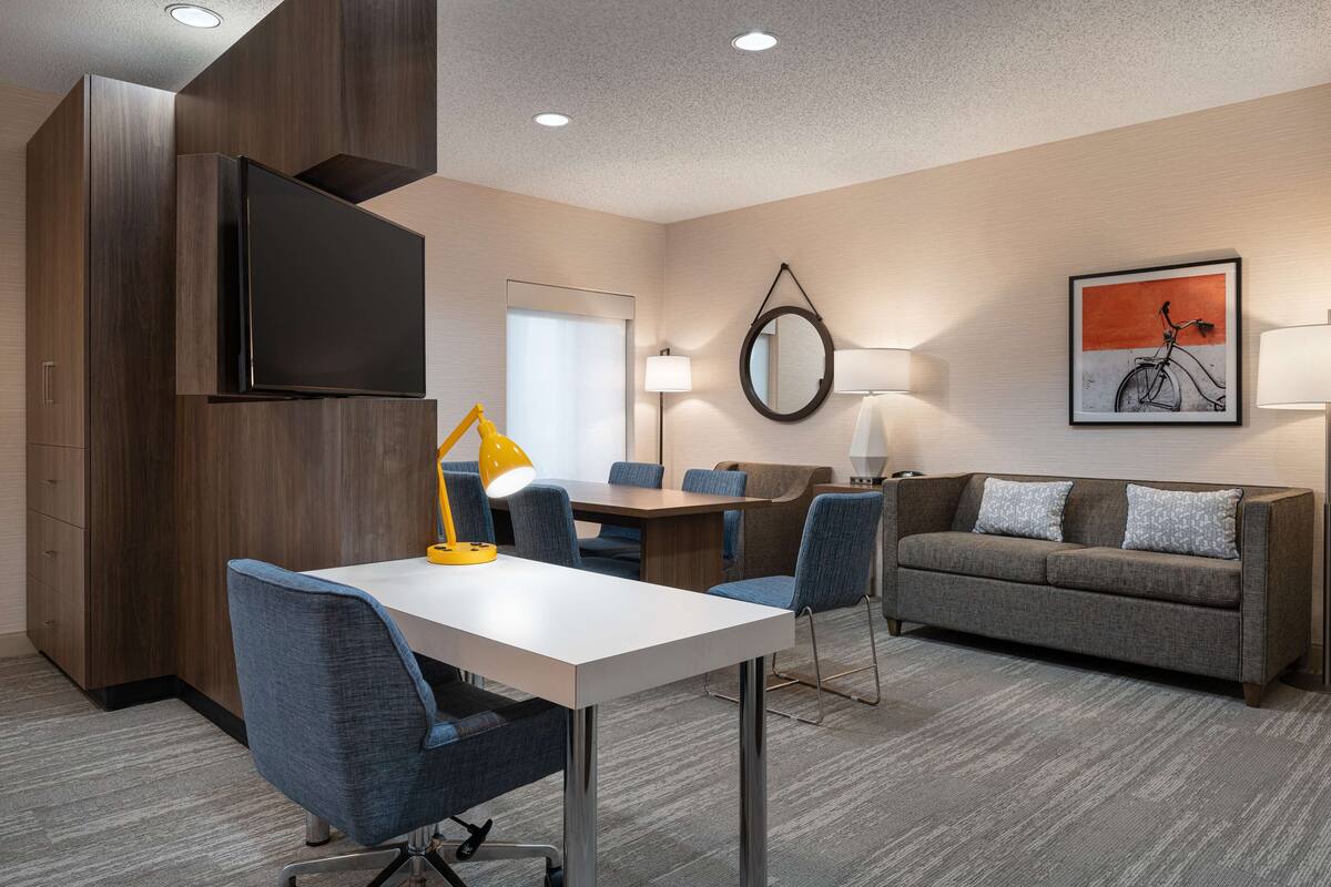A modern hotel room with a flat-screen TV, a dining table with chairs, a sofa, and a large mirror.