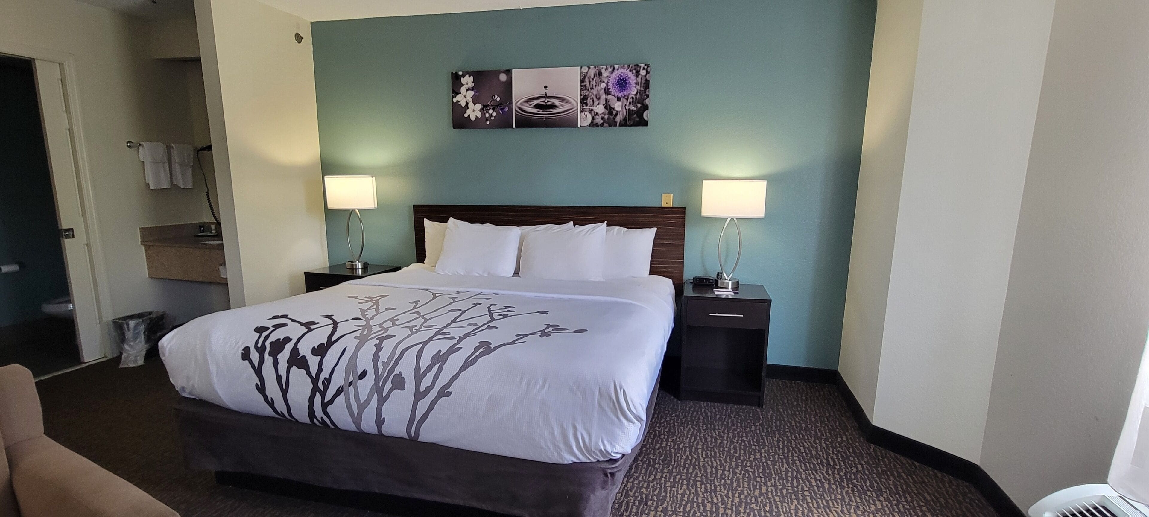 Une chambre d’hôtel avec un grand lit, deux tables de chevet avec des lampes, une salle de bain visible à travers une porte ouverte et des œuvres d’art accrochées au mur.