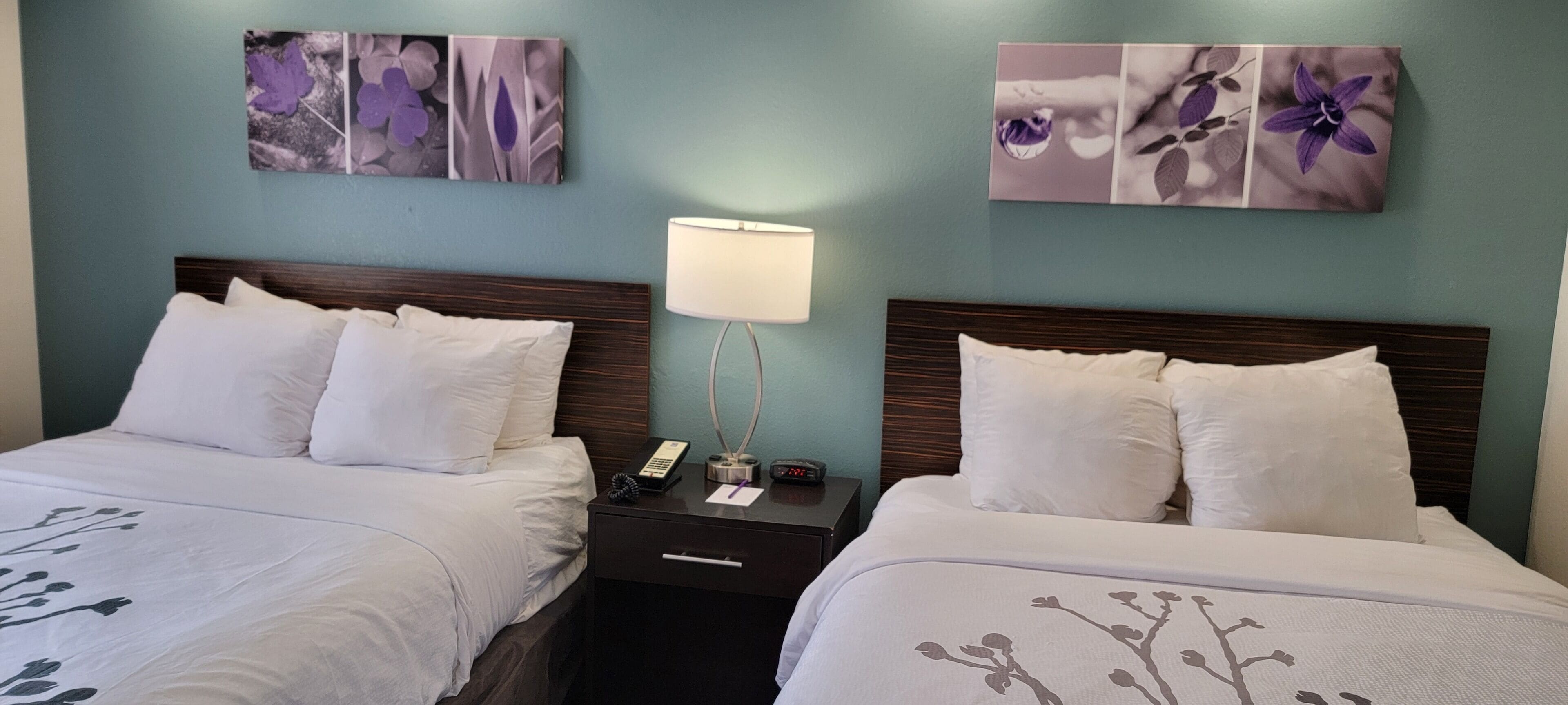Una camera d'albergo con due letti, un comodino, una lampada e decorazioni floreali alle pareti.