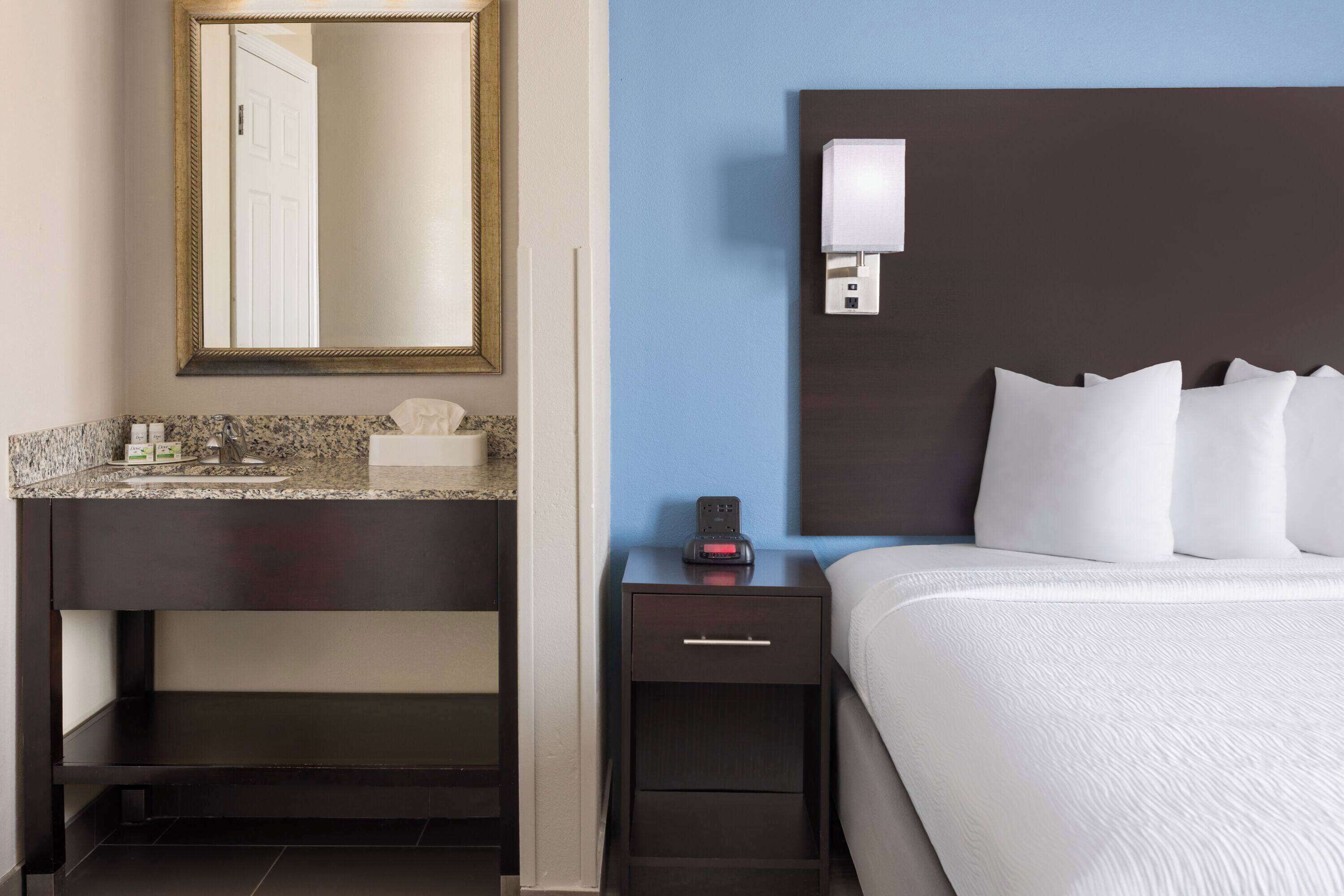 A hotel room with a dark wooden nightstand, a white bed with multiple pillows, a marble countertop, a small drawer, a wall-mounted light, and a large mirror above the countertop.
