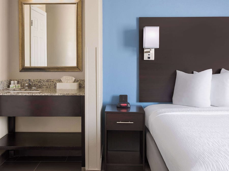 A hotel room with a dark wooden nightstand, a white bed with multiple pillows, a marble countertop, a small drawer, a wall-mounted light, and a large mirror above the countertop.