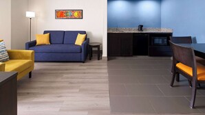 Una sala moderna con un sofá azul, un sillón amarillo y una pequeña mesa redonda. Junto a la sala se encuentra una cocina con armarios oscuros y una pared azul.