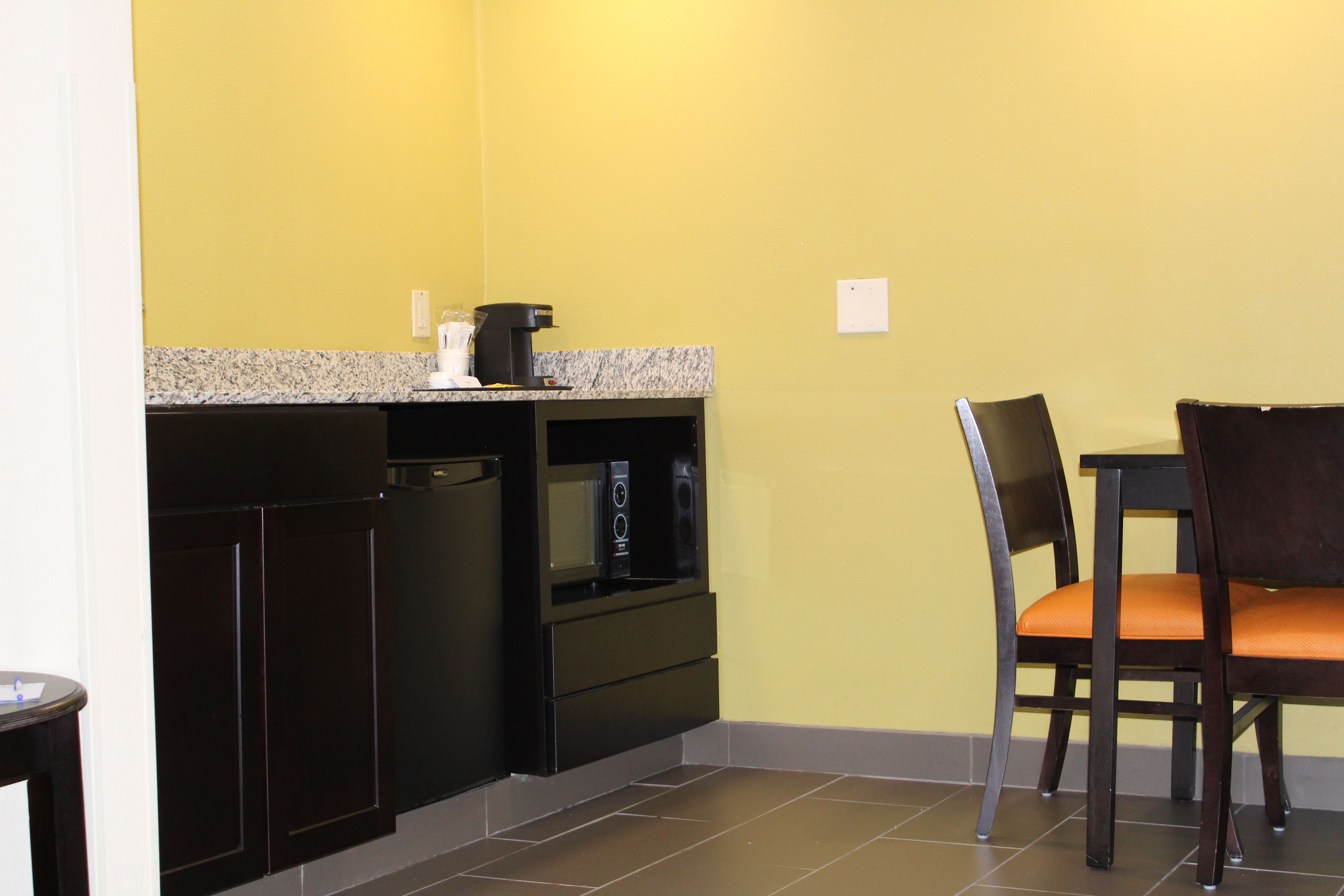 A compact kitchen with dark cabinetry, a granite countertop, and a microwave. There is a chair with an orange cushion and a small table.