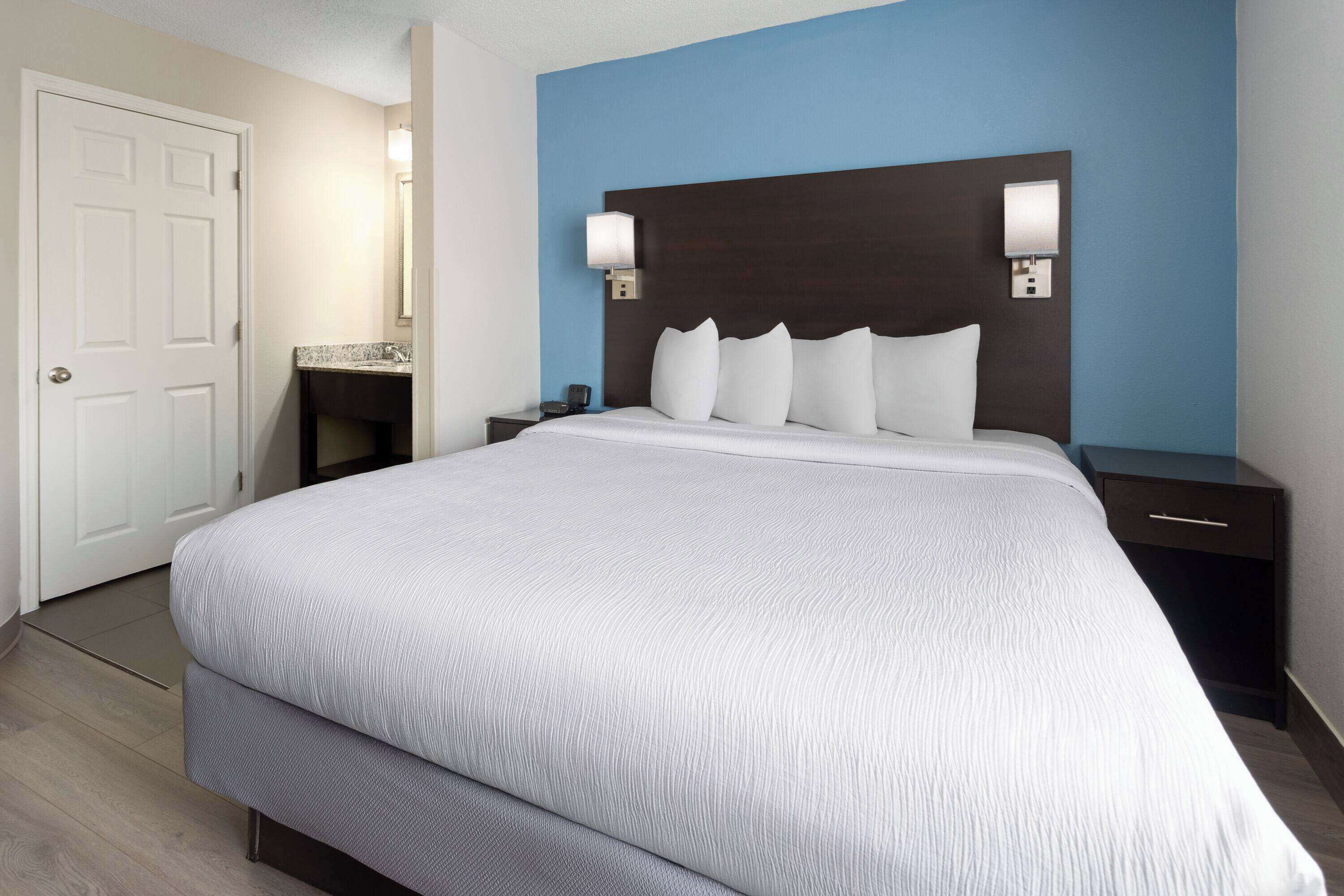A hotel room with a large bed, a nightstand, a dresser, a bathroom with a sink and mirror, and a door.