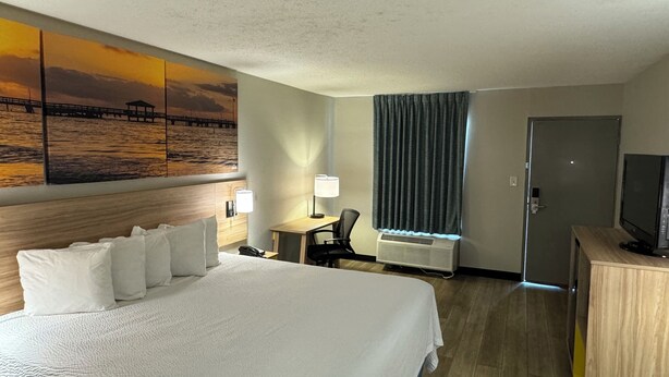 Una habitación de hotel con una cama grande, un escritorio, una silla, un televisor, un cuadro de un atardecer sobre el agua y una puerta.