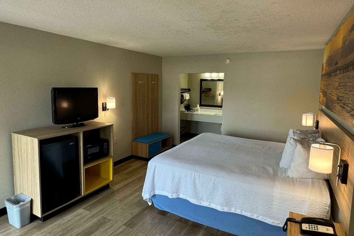 A hotel room with a bed, a TV, a desk with a computer, a mirror, and a small bathroom area.