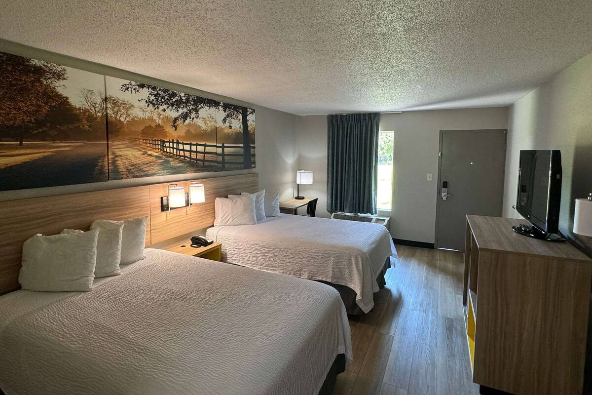 A hotel room with two beds, a large painting on the wall, a desk with a lamp, a television, and a door.