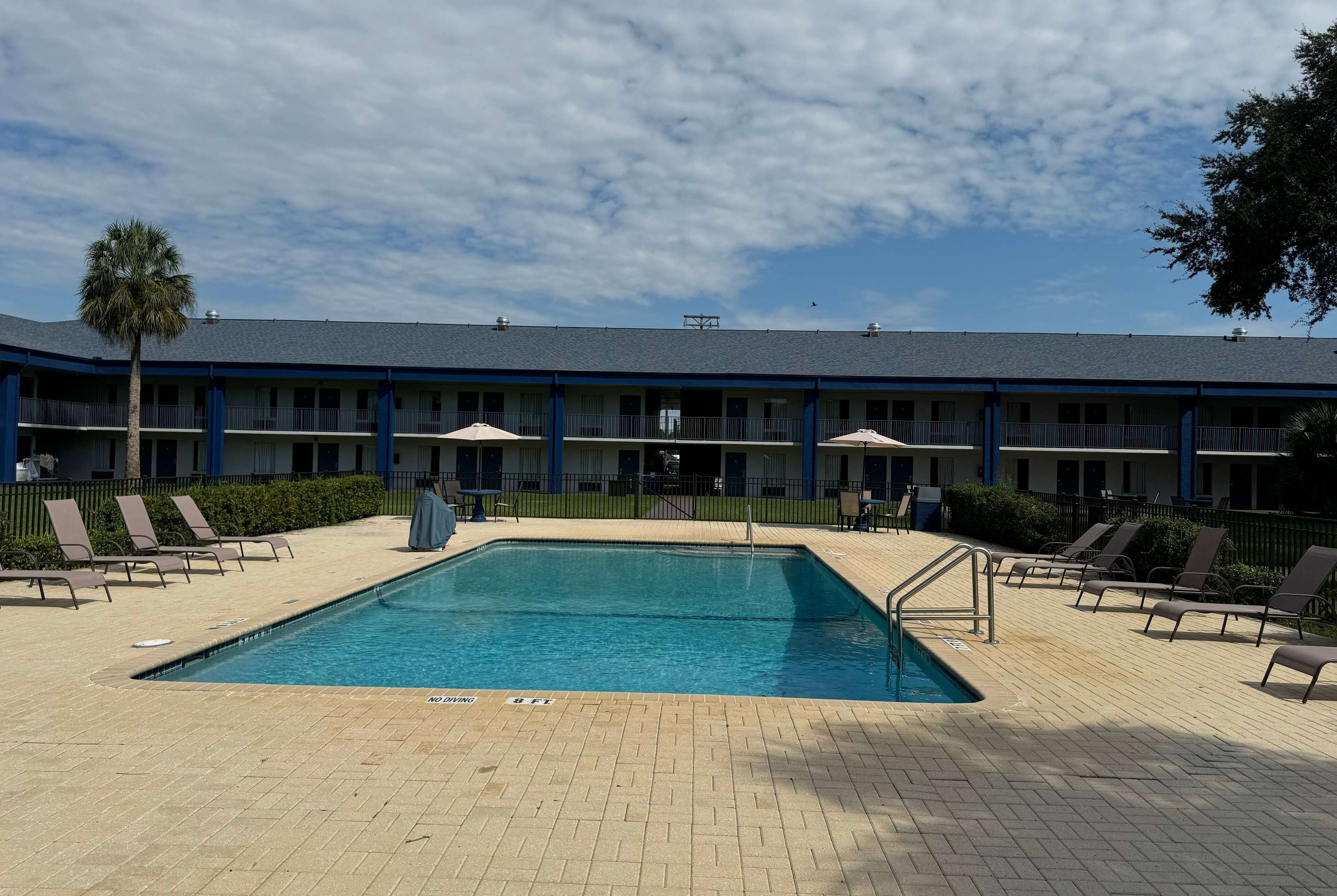Un área de piscina con sillones de descanso y sombrillas, un edificio con varios balcones y un cielo despejado.