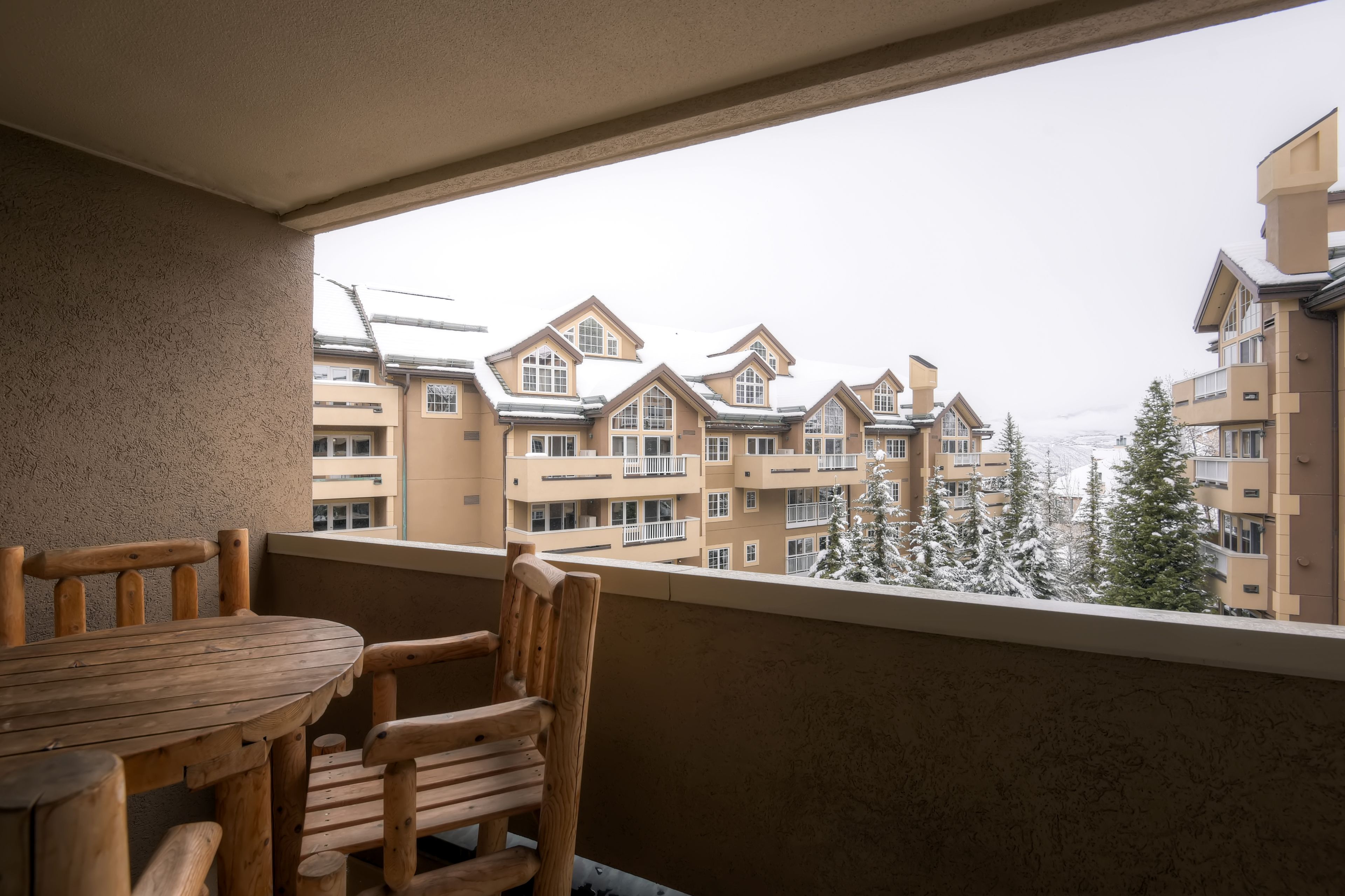 A balcony with a wooden table and chairs, overlooking a row of multi-story buildings with snow-covered trees.