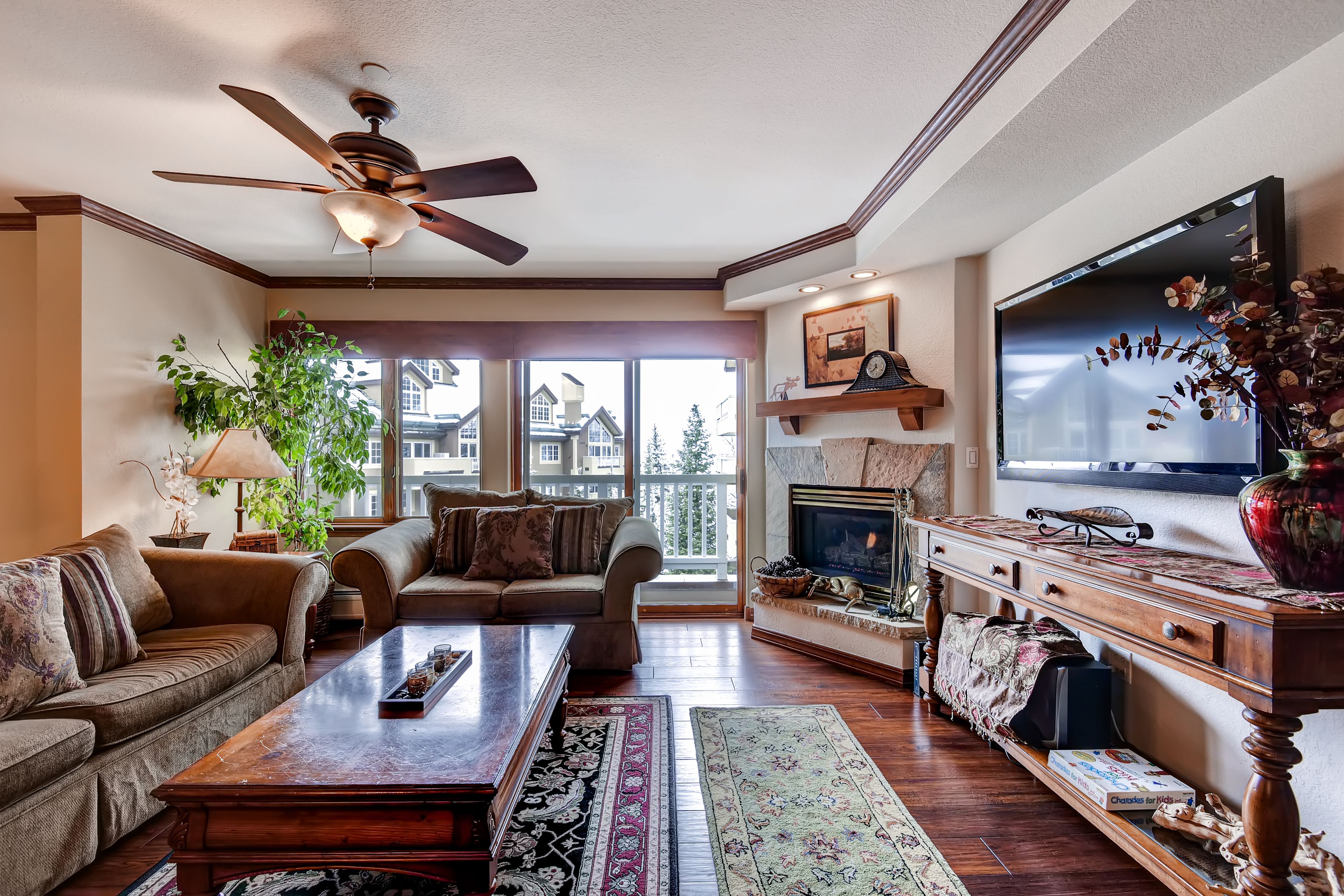 A spacious living room with a ceiling fan, a large flat-screen TV, a fireplace, and a view of the outside through large windows.