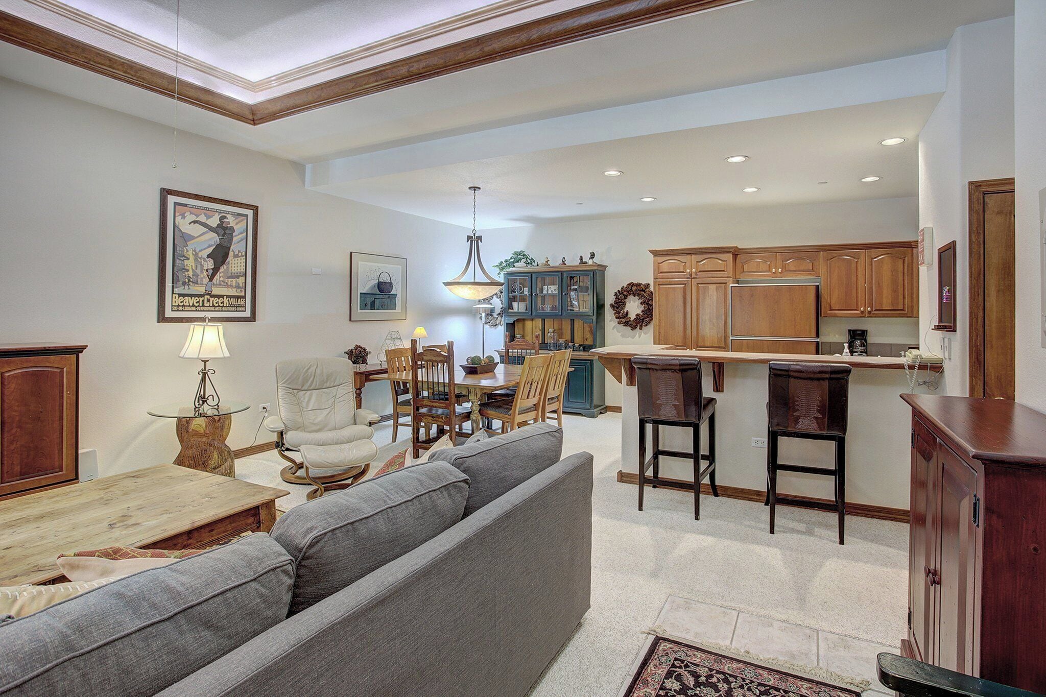 A spacious living room with a grey sofa, wooden dining table, and a kitchen area with wooden cabinets and a bar.
