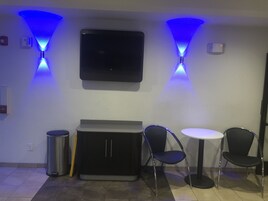 A room with a flat-screen TV mounted on the wall, two blue ceiling lights, a trash bin, a cabinet, and two chairs around a small round table.