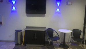Ein Zimmer mit einem an der Wand befestigten Flachbildfernseher, zwei blauen Deckenleuchten, einem Mülleimer, einem Schrank und zwei Stühlen um einen kleinen runden Tisch.