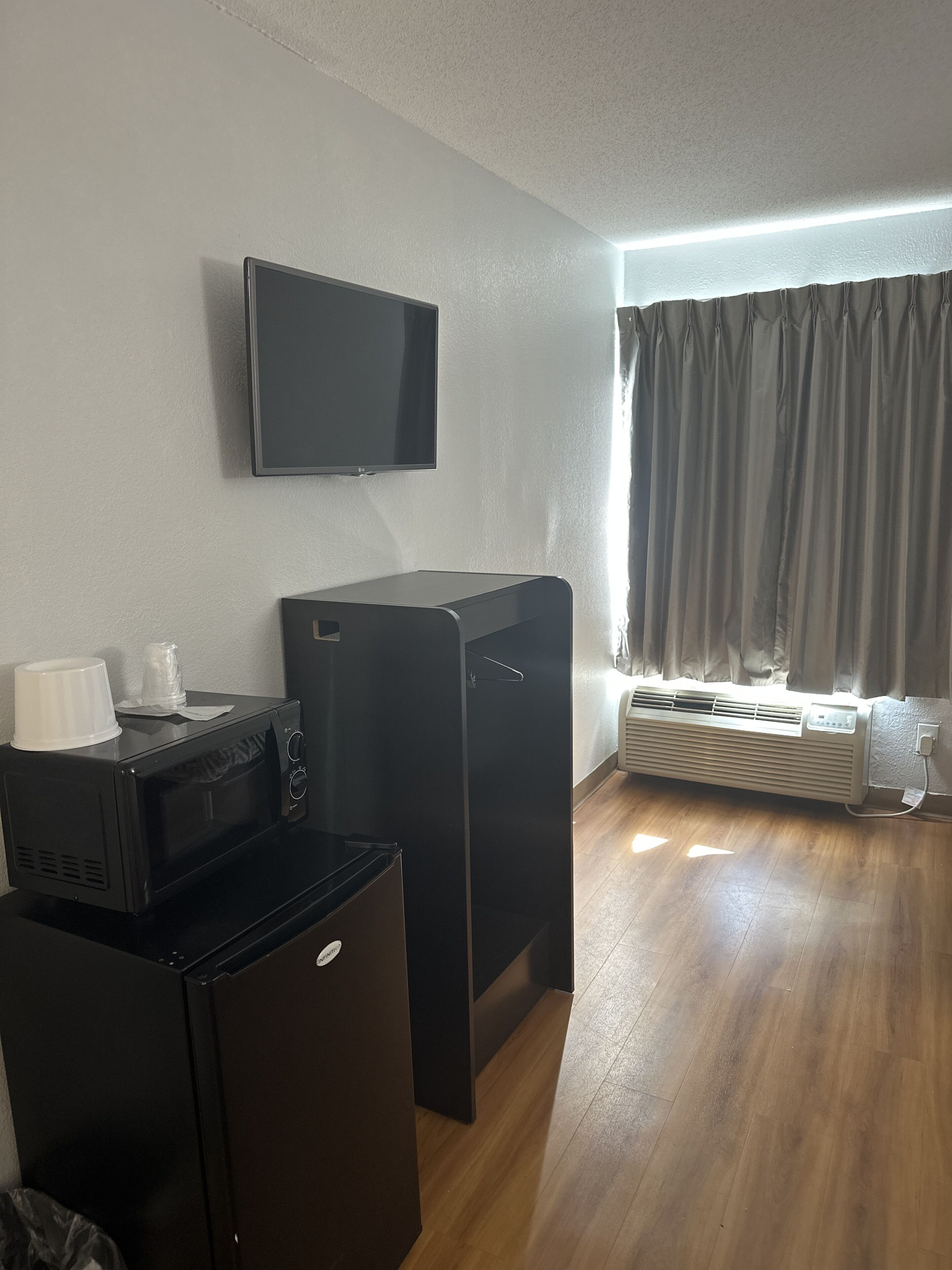 A compact room with a flat-screen TV mounted on the wall, a black refrigerator, a microwave, and a small heater near the window.